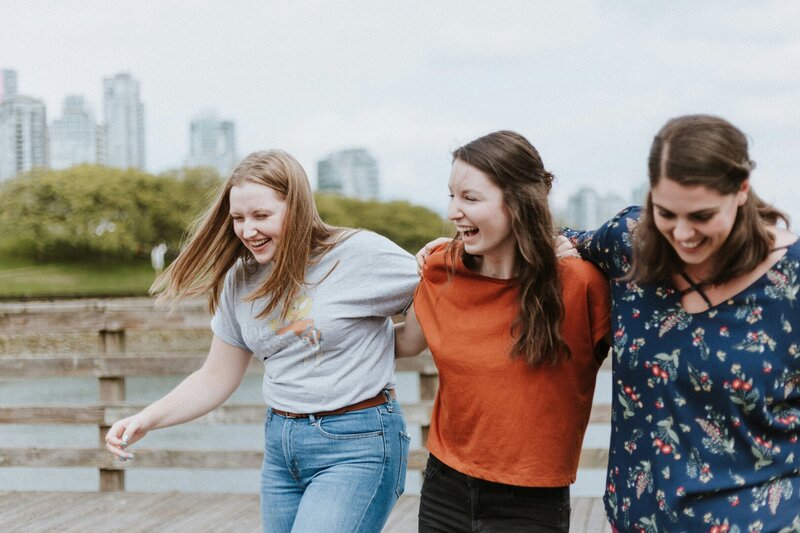 Drei junge Frauen am Wasser vor der Skyline einer Großstadt.