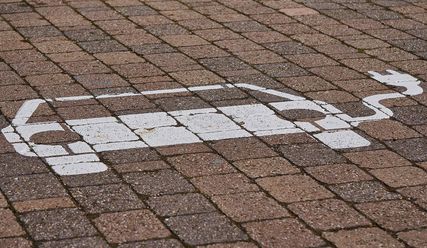 Ein weißes E-Fahrzeug als Symbol eines PKW mit Stecker und Kabel auf einem gepflasterten Parkplatz. - Copyright: Markus Distelrath auf Pixabay